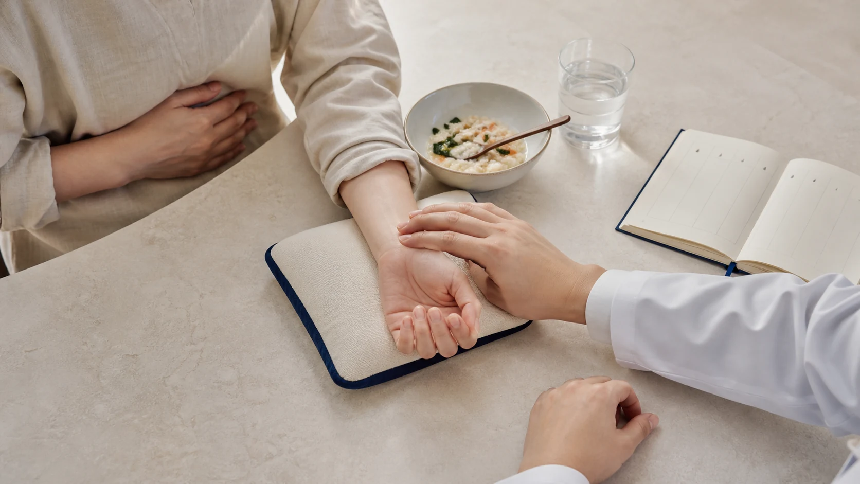 작은 식사와 증상 기록을 곁에 두고 윗배 불편을 상담하며 손목 맥을 짚는 청룡한의원 진료 장면